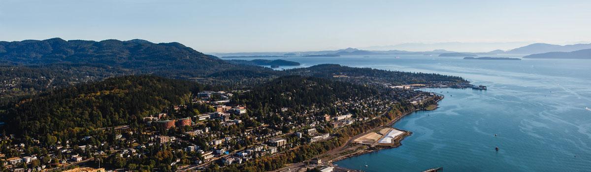 Aerial view of Western Washington University with forested hills, a bay, and distant mountains under a clear sky.